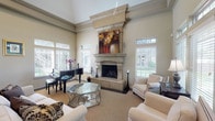 Located at 3015–3069 E 51st St in Tulsa, Oklahoma, this single-story community space is defined by its impressive architectural design and refined interior details. The centerpiece of the main gathering area is a dramatic floor-to-ceiling stone fireplace with an ornate mantel, flanked by large windows fitted with plantation shutters that allow generous natural light to fill the room. Soaring ceilings with crown molding and a ceiling fan enhance the sense of openness and grandeur throughout the space.

The main lounge area is comfortably furnished with upholstered seating arranged around a glass-top coffee table, creating an inviting setting for socializing or relaxation. A grand piano positioned near the windows adds a distinguished, elegant touch. An adjacent dining area features a large rectangular table with cushioned chairs and direct access to an outdoor patio through a glass-paneled door, offering views of the surrounding greenery. Transom windows throughout connect the dining and lounge areas visually, maintaining a cohesive, open flow.

The flooring features a neutral patterned carpet that complements the warm, taupe-toned walls. Overall, the space presents a polished, welcoming ambiance suited for gatherings, events, or everyday community use.