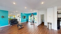 Located in Red Bank, TN, this fitness center offers a thoughtfully designed, single-floor layout that comfortably accommodates a full range of workout equipment. The space features rich hardwood-style flooring throughout, recessed lighting, and bright white walls accented by bold teal and yellow feature walls that create an energetic atmosphere. Large windows along multiple walls flood the interior with natural light and provide views of the surrounding greenery, enhancing the overall workout experience.

The main exercise area is generously proportioned, housing a comprehensive selection of strength training equipment including a multi-station cable machine, a power rack, and a full dumbbell rack with weights ranging across multiple increments. A wall-mounted flat-screen TV adds convenience for entertainment during workouts. Full-length mirrors line one wall, visually expanding the space and supporting proper form during exercise.

A secondary area is dedicated to cardio equipment, featuring treadmills and stationary bikes arranged around a striking large-format wall mural, along with a ceiling fan for added air circulation. A medicine ball rack and TRX suspension system round out the functional training options. The facility also includes a restroom and is finished with clean, modern trim work throughout, making it a complete and inviting fitness destination.