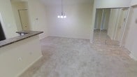 This single-story space presents a clean, neutral aesthetic throughout its open layout. The main living area features wall-to-wall plush carpeting in a warm beige tone, creating a comfortable and cohesive feel underfoot. Soft cream-colored walls reflect natural and artificial light evenly, contributing to the bright, airy atmosphere of the space.

A decorative multi-bulb chandelier serves as a focal point, suspended from the ceiling and providing warm ambient lighting to the dining or living area below. The ceiling height lends a sense of openness to the room.

Adjacent to the carpeted area, tile flooring in a neutral stone-like finish is visible, likely marking a transition into a kitchen or entryway zone. A countertop with a dark granite or laminate surface is partially visible, suggesting a kitchen or breakfast bar nearby.

Mirrored sliding closet doors are visible along one wall, adding depth and reflectivity to the space while maximizing storage potential. The overall design is understated and move-in ready, offering a versatile, well-maintained environment suited to a variety of interior styles.