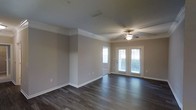 This single-floor space offers a clean, well-appointed interior defined by thoughtful architectural details and a neutral, welcoming palette. The main living area is anchored by rich, dark hardwood flooring that runs throughout, providing a striking contrast against the light-toned walls and crisp white baseboards and crown molding. The crown molding adds a refined, finished look to the space, lending an air of quality craftsmanship to the overall design. A ceiling fan with an integrated light fixture is centrally positioned, ensuring both comfort and functionality year-round. One of the standout features of this space is the set of French doors, which flood the interior with an abundance of natural light and provide a seamless connection to the outdoors. A side window further enhances the bright, airy ambiance of the room. The neutral wall tones create a versatile backdrop, making the space highly adaptable to a variety of interior styles and furnishings. The open layout allows for flexible furniture arrangements, maximizing the sense of space and flow. Overall, this space combines practical features with classic design elements to create a comfortable and inviting environment.