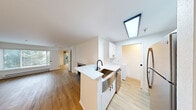 Located at 600 7th Ave in Seattle's First Hill neighborhood, this one-bedroom, two-bathroom residence offers a well-designed single-level layout. The open-concept living and kitchen area is anchored by light hardwood flooring and crisp white walls, creating a clean, modern aesthetic. The kitchen features white cabinetry, a white quartz peninsula with an undermount sink and black matte faucet, stainless steel appliances, and a skylight overhead that draws in natural light. A tile-surround fireplace serves as a focal point in the living room, which is further brightened by a large window with views of surrounding greenery. The bedroom is carpeted for comfort, outfitted with a ceiling fan, and opens directly onto a private balcony through sliding glass doors, offering outdoor access and natural light. Both bathrooms are finished in white with wood-look flooring; one features a full tub-shower combination with white tile surround and polished chrome fixtures. A dedicated laundry area adds everyday convenience. The overall palette of whites and warm wood tones throughout the unit lends a fresh, cohesive feel to the entire space.