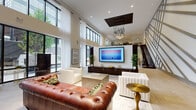 Located at 22 Sussex St in Hackensack, NJ, this contemporary amenity lounge is defined by its dramatic double-height ceilings and expansive floor-to-ceiling windows that flood the space with natural light and provide views of the surrounding greenery. The main lounge area features a rich tufted leather sofa alongside sleek upholstered seating arrangements, anchored by a large-format wall-mounted television set above a dark wood media console. A striking sculptural chandelier with cascading pendant lights serves as a focal point overhead, complemented by a dark wood ceiling accent panel. Adjacent to the lounge, a dedicated games area includes a full-size billiards table set atop a vintage-style area rug, accompanied by casual lounge seating. A bold geometric wall mural adds an artistic dimension to this zone. An open mezzanine level, accessible via a modern open-riser staircase with glass and steel railings, overlooks the main floor. The space also features a bar-height counter area with seating, a decorative gold octagonal mirror, and light tile flooring throughout. A dedicated elevator lobby with patterned tile flooring and dual elevators ensures convenient multi-floor access. The overall ambiance is upscale, social, and thoughtfully designed for both relaxation and recreation.