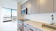 Located at 632 Newark Avenue in Jersey City, NJ, this modern unit offers a thoughtfully designed layout that maximizes every square foot. The living room is bathed in natural light from expansive floor-to-ceiling windows that frame impressive panoramic city skyline views, creating a bright and airy atmosphere throughout. Light-toned hardwood flooring runs seamlessly across the living spaces, complementing the clean, minimalist aesthetic of the interior.

The kitchen is outfitted with sleek gray cabinetry extending to the ceiling for ample storage, a white subway tile backsplash, light quartz countertops, and stainless steel appliances including a built-in over-the-range microwave. The space flows efficiently into the main living area, making it ideal for everyday living.

A well-lit hallway connects the rooms, lined with louvered closet doors offering convenient storage access. Two dedicated closets provide generous built-in storage. The bathroom features tile flooring and a clean, functional design. In-unit laundry adds a practical convenience. Crisp white walls throughout, recessed lighting in the hallway, and a consistent modern finish give the entire unit a polished, move-in ready feel.