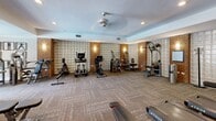 Located at 9030 Lorton Station Blvd in Lorton, Virginia, this fitness center offers a thoughtfully designed exercise space that balances functionality with refined aesthetics. The room is defined by its distinctive architectural character, featuring exposed brick columns interspersed with translucent glass block wall panels that diffuse natural light softly throughout the interior. Recessed ceiling lights and wall-mounted sconces provide warm, even illumination, while a ceiling fan contributes to a comfortable workout environment.

The floor is covered in durable, patterned carpet tiles that add texture and warmth to the space. Full-length mirrors line one wall, visually expanding the room and allowing users to monitor their form during exercise. A window along one wall brings in additional natural light and a connection to the outdoors.

The facility is comprehensively equipped with a broad selection of cardio machines, including treadmills, ellipticals, stair climbers, and stationary bikes. The strength training area features multi-functional cable machines, a dedicated free weights section with dumbbells and weight benches, and various resistance machines. The layout is spacious and well-organized, allowing for comfortable movement between stations. Overall, the fitness center presents a polished, well-maintained environment suited for a full-range workout experience.