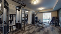 This single-floor fitness and aquatic center is thoughtfully designed to support a comprehensive wellness experience. The main workout area is generously proportioned, featuring a full suite of Life Fitness strength training equipment, including cable machines, a lat pulldown station, and a weight bench. White decorative columns, wall-length mirrors, and patterned carpet tiles in muted gray tones give the space a polished, modern aesthetic. Warm flush-mount drum lighting provides consistent illumination throughout. A dedicated cardio room houses multiple treadmills and an elliptical trainer, accompanied by mirrors and a window that connects visually to the pool area. Sliding barn-style doors open into a versatile studio space finished with light hardwood-style flooring, a ballet barre, and an assortment of free weights and exercise balls, making it suitable for stretching, yoga, or group classes. The indoor aquatic area is expansive, featuring a large lap pool surrounded by a wide concrete deck and rows of lounge chairs. Floor-to-ceiling windows along one wall flood the pool deck with natural light. The facility's layout allows seamless transitions between all areas, creating a cohesive and functional environment for a full range of fitness activities.