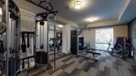 This single-floor fitness and aquatic center is thoughtfully designed to support a comprehensive wellness experience. The main workout area is generously proportioned, featuring a full suite of Life Fitness strength training equipment, including cable machines, a lat pulldown station, and a weight bench. White decorative columns, wall-length mirrors, and patterned carpet tiles in muted gray tones give the space a polished, modern aesthetic. Warm flush-mount drum lighting provides consistent illumination throughout. A dedicated cardio room houses multiple treadmills and an elliptical trainer, accompanied by mirrors and a window that connects visually to the pool area. Sliding barn-style doors open into a versatile studio space finished with light hardwood-style flooring, a ballet barre, and an assortment of free weights and exercise balls, making it suitable for stretching, yoga, or group classes. The indoor aquatic area is expansive, featuring a large lap pool surrounded by a wide concrete deck and rows of lounge chairs. Floor-to-ceiling windows along one wall flood the pool deck with natural light. The facility's layout allows seamless transitions between all areas, creating a cohesive and functional environment for a full range of fitness activities.