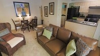 This single-story residence features an open-concept design that seamlessly integrates the living, dining, and kitchen areas into one cohesive space. The living area is anchored by a rich brown leather loveseat accented with decorative green and patterned throw pillows, complemented by a glass-top coffee table. Neutral carpeting runs throughout, contributing to a warm and inviting ambiance. Decorative bamboo stalks and framed botanical wall art add natural, organic touches to the space.

Adjacent to the living area, the dining space accommodates a dark wood table and matching chairs, ideal for everyday meals. A softly lit lamp on the dining table enhances the room's cozy atmosphere.

The kitchen is partially visible beyond a dark countertop peninsula, revealing stainless steel appliances including a full-size refrigerator. White cabinetry and light walls keep the kitchen feeling bright and functional. A glimpse of an additional room beyond the kitchen suggests further living space. Overall, the layout is efficient and well-appointed, with a consistent color palette of earth tones, greens, and neutrals creating a harmonious, livable environment.