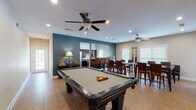 This single-story clubhouse in Atoka, Tennessee, is designed to serve as a versatile community gathering space, offering a well-rounded combination of recreation, dining, and relaxation areas. The layout flows openly between zones, all finished with neutral-toned tile flooring throughout and ceiling fans with integrated lighting in each area.

The centerpiece of the main gathering room is a full-size pool table, positioned beneath recessed lighting and ceiling fans that keep the space comfortable. The room transitions naturally into a dining area furnished with multiple high-top tables and dark wood bar-height chairs, providing generous seating capacity for group events or casual gatherings. Windows along the walls allow natural light to fill the space, and a wall-mounted cue rack keeps billiards equipment neatly organized nearby.

The kitchen is well-appointed with rich, dark wood cabinetry, a stainless steel French-door refrigerator, a dishwasher, microwave, and a built-in bar-style counter with seating. This setup makes the space practical for catering or community events.

A secondary lounge area features a sofa, a wall-mounted television, and a fireplace, accessible through French doors, offering a quieter retreat within the clubhouse. The overall ambiance is welcoming and functional, suited for a wide range of community activities.