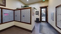 Located at 3290 Dewar Dr in Rock Springs, Wyoming, this dedicated mail room serves as a functional and well-appointed communal space. The room is organized around multiple banks of individual, numbered metal mailboxes finished in a neutral gray, set within dark wood-trimmed frames that lend a polished, cohesive aesthetic to the space. The mailbox units are arranged along the walls and on a central partition, maximizing accessibility and capacity within the compact footprint.

The interior features warm beige walls, dark wood baseboards, and durable concrete flooring complemented by entry mats near the door. A glass-paneled exterior door allows natural light to filter into the room, providing a bright and welcoming atmosphere. A bulletin board is mounted near the mailboxes, offering residents a place for community notices and postings.

Decorative touches include potted greenery and trailing ivy plants displayed atop the mailbox partitions, adding a lively, natural element to the utilitarian setting. Framed artwork on the walls further enhances the room's inviting character. Overhead recessed lighting ensures the space is well-lit throughout. A small waste bin near the entrance provides added convenience. Overall, the mail room balances practical function with thoughtful design details, creating a clean and comfortable environment for daily use.