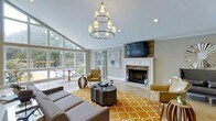 This well-appointed single-floor space makes a striking impression with its open and airy living area. Vaulted ceilings rise dramatically overhead, accentuated by a tiered chandelier with warm-toned shades that serves as a central design focal point. Large, floor-to-ceiling windows flood the interior with natural light, offering views of lush, mature greenery outside and creating a seamless connection between the indoors and the surrounding landscape.

The living area is anchored by a classic stone fireplace with a crisp white mantel, providing both a functional hearth and a refined architectural feature. A wall-mounted flat-screen television above the mantel integrates modern convenience into the traditional design. The space is thoughtfully furnished with a sectional sofa, accent chairs in warm earth tones, and a bold amber-and-white patterned area rug that adds vibrancy and warmth to the light neutral palette of the walls and flooring.

A casual dining or reading nook is nestled near the windows, taking full advantage of the natural light. Crown molding and recessed lighting complement the chandelier, adding polish throughout. The overall ambiance is inviting and sophisticated, blending contemporary furnishings with timeless architectural details to create a comfortable and visually engaging environment.