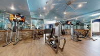 Located at 3491 SW 42nd St in Ocala, Florida, this fitness facility is a single-floor space designed to deliver a comprehensive and visually engaging workout experience. The facility is divided into several distinct training zones, each thoughtfully finished and equipped.

The primary strength and machine training area is distinguished by a soaring vaulted ceiling adorned with a sky-and-clouds mural, complemented by floor-to-ceiling mirrored walls that amplify the sense of space. Large ceiling fans provide air circulation, and recessed lighting ensures the area is well-lit. The lower walls feature a striking combination of natural stone veneer and wood shiplap paneling, grounding the airy ceiling design with a rustic, warm aesthetic. A variety of Life Fitness selectorized machines and cable stations are arranged throughout this zone, alongside wall-mounted televisions.

A dedicated cardio area showcases the same navy blue shiplap accent walls with motivational phrases like "Don't Wish For It… Work For It," paired with stone wainscoting. This section houses treadmills, stationary bikes, elliptical machines, and medicine balls, with natural light filtering through arched windows.

A separate strength alcove features rubber flooring and a Life Fitness Smith machine with a bench, ideal for free-weight training. Throughout the entire facility, light hardwood-style flooring unifies the space, creating a clean, modern foundation that complements the facility's distinctive and motivating design aesthetic.