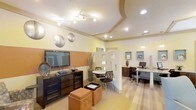This single-floor space is thoughtfully designed to accommodate multiple functions within an open, flowing layout. The interior is divided into distinct zones using a decorative partial-height room divider featuring geometric cutout windows, which maintains visual connectivity while subtly separating areas. Warm yellow and terracotta accent walls contribute to the welcoming ambiance throughout.

The lounge area anchors one side of the space, featuring a wall-mounted flat-screen television atop a dark media console with ample storage. Decorative metallic disc wall art adds an artistic focal point, while a pair of rust-colored upholstered ottomans provide casual seating. Track lighting along the ceiling illuminates this zone effectively.

Beyond the divider, a compact dining area with a round table and modern black chairs offers a casual seating option. Adjacent to this, a dedicated workspace features a granite-topped desk, ergonomic seating, a desktop computer, and built-in display niches with teal backing, adding a pop of color. Recessed lighting and pendant fixtures ensure the work area is well-lit. The flooring transitions subtly across zones, enhancing the spatial definition of each area.