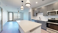 This thoughtfully designed single-floor residence offers a clean, modern aesthetic throughout its well-organized layout. The open-concept kitchen serves as a central feature, showcasing gray flat-front cabinetry, a geometric hexagonal tile backsplash, stainless steel appliances including a French door refrigerator, range, microwave, and dishwasher, and a large quartz-topped island illuminated by stylish pendant lights. Light wood-look flooring runs throughout the main living areas, lending warmth and continuity.

The living area is bright and airy, benefiting from natural light through blinds-fitted windows and a ceiling fan overhead. A glazed door provides additional access and light. The bedroom is a comfortable, carpeted retreat with a ceiling fan, a large window for natural light, and access to a walk-in closet equipped with wire shelving and ample storage.

The bathroom features light wood-look flooring, a full soaking tub with tile surround, a vanity with storage, and a clean, minimal design. A hallway connects the rooms efficiently, completing the functional floor plan. Overall, the space delivers a fresh, contemporary living environment with quality finishes and practical design throughout.