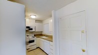 This single-floor space showcases a compact yet functional kitchen that forms a central feature of the unit. The kitchen is designed in an L-shaped layout, maximizing the use of available space while maintaining clear workflow between key areas. White cabinetry lines the upper and lower walls, providing ample storage and contributing to a clean, bright aesthetic throughout the space.

The kitchen is equipped with a full suite of white appliances, including an electric range with coil burners, a built-in over-the-range microwave, a dishwasher, and a full-size refrigerator. A single-basin sink is positioned at the corner of the L-shaped counter, fitted with a standard faucet and bordered by dark-toned laminate countertops that offer a subtle contrast to the white cabinetry. Vertical blinds on the window above the sink allow for adjustable natural light.

Light tile flooring runs through the kitchen, adding to the overall brightness of the space. The ceiling features a textured popcorn finish with a flush-mount light fixture providing overhead illumination. A door near the kitchen entry provides access to an adjacent area, contributing to the unit's practical flow and layout.