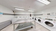 This shared laundry facility at 3122 Hurontario St in Mississauga, Ontario is a practical, efficiently organized space designed to accommodate high-volume use. The room is finished with clean white wall tiles accented by a horizontal band of grey tiles, creating a crisp and hygienic aesthetic throughout. Light-toned floor tiles complement the bright interior, which is illuminated by fluorescent ceiling fixtures that ensure consistent, even lighting across the entire space.

A row of front-load washing machines lines one wall, each equipped with individual digital control panels for ease of use. Opposite the washers, a corresponding row of top-load dryers provides ample capacity for simultaneous loads. Two large stainless steel folding tables are centrally positioned, offering generous workspace for sorting and folding laundry. A freestanding stainless steel utility sink is also present, adding functional versatility to the room.

Additional amenities include a coin or card-operated payment station, informational signage, and a security camera for resident safety. A grey steel entry door with a frosted glass panel provides secure access to the facility. The layout is thoughtfully arranged to allow comfortable circulation around all equipment, making this laundry room a fully functional and well-maintained amenity.