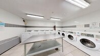 This shared laundry facility at 3122 Hurontario St in Mississauga, Ontario is a practical, efficiently organized space designed to accommodate high-volume use. The room is finished with clean white wall tiles accented by a horizontal band of grey tiles, creating a crisp and hygienic aesthetic throughout. Light-toned floor tiles complement the bright interior, which is illuminated by fluorescent ceiling fixtures that ensure consistent, even lighting across the entire space.

A row of front-load washing machines lines one wall, each equipped with individual digital control panels for ease of use. Opposite the washers, a corresponding row of top-load dryers provides ample capacity for simultaneous loads. Two large stainless steel folding tables are centrally positioned, offering generous workspace for sorting and folding laundry. A freestanding stainless steel utility sink is also present, adding functional versatility to the room.

Additional amenities include a coin or card-operated payment station, informational signage, and a security camera for resident safety. A grey steel entry door with a frosted glass panel provides secure access to the facility. The layout is thoughtfully arranged to allow comfortable circulation around all equipment, making this laundry room a fully functional and well-maintained amenity.