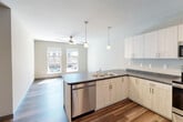 This thoughtfully designed 2-bedroom, 1-bathroom unit offers a clean, modern aesthetic across a single floor. The open-concept kitchen and living area serves as the heart of the space, featuring light wood-grain cabinetry, dark contrasting countertops, a stainless steel dishwasher and range, a double-basin sink, and a peninsula that defines the kitchen zone. Pendant lighting and a ceiling fan with light fixture enhance both function and style, while large windows flood the living area with natural light and provide views of the surrounding streetscape. Durable wood-look flooring runs throughout the main living areas, adding warmth and visual continuity. The two bedrooms are carpeted for comfort, each benefiting from natural light through generously sized windows. The primary bedroom offers a comfortable footprint, while the second bedroom features the same neutral palette and hardwood-style flooring. The bathroom is finished with a clean white vanity, undermount sink, chrome fixtures, and a large mirror, accompanied by convenient closet access. Two additional closets throughout the unit provide ample storage. A hallway connects the private rooms seamlessly to the main living areas, completing this well-organized and contemporary residential layout.