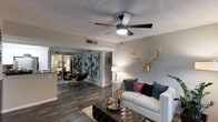 This well-appointed single-floor residence offers a thoughtfully designed open-concept layout that connects the living and dining areas in a seamless, functional arrangement. The living room is anchored by dark hardwood-style flooring, a white sofa accented with rich jewel-toned throw pillows, and a tufted leather ottoman that serves as both a coffee table and a centerpiece. A statement gold antler mount adorns the accent wall, adding a bold, eclectic touch to the otherwise contemporary aesthetic. A ceiling fan with an integrated light fixture provides both comfort and ambiance overhead, while a textured popcorn ceiling adds a classic residential character. Decorative geometric wall art creates a visual divider between the living and dining spaces without sacrificing the open feel of the layout. The dining area accommodates a round table surrounded by sleek black chairs, complemented by a mirrored wall feature that reflects light and creates a sense of expanded space. A pass-through or partial wall connects the dining area to what appears to be a kitchen beyond. Neutral gray walls throughout the space provide a versatile backdrop that ties the varied design elements together cohesively, resulting in an inviting and personality-filled interior.