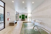 Located at 1308 Lorna St in Charlotte, NC, this spa facility offers a tranquil, well-appointed retreat designed with relaxation and wellness in mind. The centerpiece of the space is a recessed hydrotherapy pool fed by cascading waterfall jets built into a textured pebble-stone accent wall, creating both a visual and auditory sense of calm. The pool is accessible via sleek stainless steel handrails and is surrounded by travertine tile flooring that extends throughout the room, lending a warm, natural aesthetic. Horizontal bands of mosaic pebble tile adorn the walls, adding tactile depth and visual interest to the overall design. Recessed ceiling lighting illuminates the space evenly, while large glass-panel doors and windows along the perimeter allow natural light to flow in freely. Two vibrant living green wall panels serve as a bold focal point at one end of the room, complemented by a potted tropical plant and a minimalist bench that reinforce the spa's organic, wellness-oriented atmosphere. A wall-mounted split HVAC unit ensures year-round climate comfort. An adjacent area, visible through full-height glass partitions, features secured lockers, providing practical amenities for guests. The overall ambiance is clean, modern, and restorative, making this space a compelling wellness amenity.