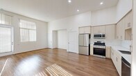 This thoughtfully designed single-floor unit offers an efficient and open layout that maximizes every square foot. The combined kitchen and living area forms the heart of the space, characterized by soaring ceilings and an abundance of natural light streaming through large windows fitted with vertical blinds. Warm-toned hardwood flooring runs throughout, adding a sense of continuity and comfort to the open floor plan.

The kitchen is well-appointed with a full suite of stainless steel appliances, including a French door refrigerator, gas range, built-in microwave, and dishwasher. Light wood cabinetry extends floor-to-ceiling, providing generous storage, while a white tile backsplash and a clean countertop with an undermount sink complete the functional and modern aesthetic. Recessed lighting above ensures the space remains bright and inviting.

The bathroom is finished to a high standard, featuring marble-patterned tile surrounds in the tub-shower combination, light wood vanity cabinetry, a large mirror, built-in open shelving, and neutral floor tile — all contributing to a clean, spa-like atmosphere.

The living area benefits from a private entry door and expansive windows that flood the room with daylight, while the crisp white walls and high ceilings create an airy, open ambiance throughout.