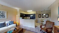 This single-floor residence offers a thoughtfully designed open-concept layout that seamlessly connects the living, kitchen, and dining areas. The living room features light wood-look tile flooring, warm beige walls, and windows that invite natural light, creating a relaxed coastal ambiance accented by nautical-themed décor. The kitchen is a standout feature, boasting dark espresso cabinetry, granite countertops, a full suite of black appliances including a range, microwave, and refrigerator, and a peninsula that opens to the main living space — ideal for both everyday use and entertaining. Adjacent to the kitchen, the dining area accommodates a round dining table beneath a pendant light fixture, with decorative wall mirrors adding depth to the space. Both bedrooms are carpeted and generously sized, each with large windows that allow ample natural light and views of the surrounding greenery. The rooms offer closet storage and ceiling fans for comfort. The bathroom mirrors the kitchen's design aesthetic, featuring matching dark cabinetry, granite countertops, a wide vanity mirror, and elegant tile flooring. Throughout the residence, the warm neutral palette and consistent finishes create a cohesive, inviting atmosphere.