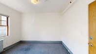 This single-floor space presents a straightforward, functional layout with a clean and neutral aesthetic throughout. The rooms are finished with white walls and blue-grey carpeting, creating a calm and versatile environment. Consistent grey baseboards provide a subtle contrast to the bright wall surfaces, lending a cohesive look across the unit.

Several rooms benefit from multi-pane windows with warm wood frames, allowing natural light to pour in and cast bright patches across the carpeted floors. Each windowed room is equipped with a cast-iron radiator, providing reliable, traditional heating. Ceiling fixtures offer additional overhead lighting where windows are absent.

The layout includes multiple rooms of varying sizes, some featuring structural details such as exposed ceiling beams and built-in utility panels. A fire suppression sprinkler system is present, adding a practical safety element throughout.

The bathroom is bright and simply appointed, featuring white walls, vinyl flooring, a wall-mounted sink, a medicine cabinet mirror, a towel bar, and dual multi-pane windows that bring in generous natural light. A cast-iron radiator in the bathroom ensures year-round comfort.

Overall, the space offers a clean, move-in-ready environment with consistent finishes, ample natural light, and practical infrastructure suited for a range of uses.