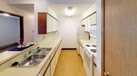 This two-bedroom, one-bathroom unit is thoughtfully laid out across a single floor, making efficient use of its space. The living room serves as the central gathering area, featuring a vaulted ceiling that adds a sense of openness and architectural character. Track lighting and a ceiling fan fixture complement the room, while a large sliding glass door leads to an outdoor balcony, filling the space with natural light. Carpet and hard-surface flooring meet at the transition between the living and kitchen areas.

The galley-style kitchen is well-equipped with ample white cabinetry along both walls, a double stainless steel sink, laminate countertops, and white appliances including a refrigerator, electric range, and built-in microwave. Light wood-tone flooring runs throughout the kitchen, lending a clean and practical finish.

Both bedrooms are carpeted and feature windows with blinds, baseboard heating, and flush-mount ceiling light fixtures, offering comfortable and functional spaces. A walk-in closet with wire shelving provides generous storage for one of the bedrooms.

The full bathroom includes a wide vanity with a large mirror, a multi-bulb light bar, wood-toned cabinetry, hard-surface flooring, and a shower-tub combination enclosed by a curtain. A textured ceiling runs consistently throughout the unit.