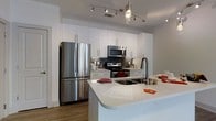 This single-floor residence showcases a thoughtfully designed, open-concept kitchen that serves as the centerpiece of the space. The kitchen is outfitted with a full suite of stainless steel appliances, including a French door refrigerator, an electric range with microwave above, and a dishwasher, all complemented by a classic subway tile backsplash in a cool gray tone. Crisp white shaker-style cabinetry extends floor to ceiling, providing generous storage while lending a clean, contemporary aesthetic. The expansive island anchors the kitchen, featuring a double stainless steel sink with a modern gooseneck faucet and a smooth white quartz countertop that offers ample workspace and casual seating potential. Overhead, a stylish curved track lighting fixture provides focused, adjustable illumination throughout the kitchen, while a pendant light adds an elegant accent above the island. Warm-toned hardwood-style flooring runs throughout, adding depth and contrast to the predominantly white and gray palette. The neutral gray walls and open sightlines create an airy, welcoming atmosphere, making this kitchen as functional as it is visually appealing.