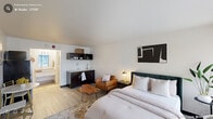 Located on Carolina Ave in Durham, NC, this efficient studio-style unit offers a well-designed single-floor layout with one bedroom and one bathroom. The main living area serves as the focal point of the space, featuring light wood-style flooring, smooth white walls, and clean-lined architecture that creates a bright, open ambiance. The bedroom area is generously proportioned relative to the overall footprint, accommodating a full-size bed with an upholstered headboard, a leather accent chair, and a live-edge wood coffee table that adds a natural, organic touch to the modern aesthetic. A wall-mounted flat-screen TV and a tall leaning mirror enhance both functionality and the sense of space. A compact dining area with a round marble-top table and gold-accented chairs provides a dedicated spot for meals. The kitchenette is fitted with dark cabinetry, open shelving, a sink, a countertop, a stainless steel refrigerator, and a microwave — offering essential cooking and storage convenience. The bathroom features a granite-topped vanity with an undermount sink and a separate shower stall with light tile flooring. Throughout the unit, the cohesive design palette of whites, neutrals, and warm wood tones creates a modern yet inviting atmosphere.