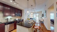 This single-floor residence offers a thoughtfully designed open-concept layout that seamlessly connects the kitchen, dining, and living areas. The kitchen is a standout feature, showcasing rich dark-cherry cabinetry with under-cabinet lighting, a mosaic tile backsplash, dark granite countertops, and a full suite of stainless steel appliances including a gas range, built-in microwave, dishwasher, and French door refrigerator. A peninsula with bar seating provides additional prep space and a casual dining option, while a dedicated dining area nearby accommodates a glass-top table with easy access to the kitchen. The living room flows naturally from the kitchen and is anchored by warm hardwood flooring and a ceiling fan. Large sliding glass doors open to a private balcony, allowing natural light to fill the space and providing direct outdoor access. The bedroom is carpeted for comfort and features a ceiling fan, a large window with sheer drapery, and ample room for a full bedroom suite. The bathroom is finished with neutral tile flooring, a granite-topped vanity, and a full soaking tub with a shower. Track lighting and pendant fixtures throughout the residence provide well-distributed illumination, contributing to the overall polished and contemporary ambiance of the home.