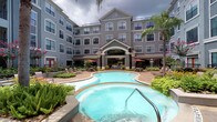 This amenity space is part of a multi-story residential community distinguished by its classic architectural design, featuring a symmetrical gray facade, arched entryways with a copper-toned roof accent, and lush tropical landscaping. At the heart of the outdoor area lies a freeform pool with a connected spa, surrounded by a decorative paved deck, lounge seating, and shade umbrellas — creating a resort-like atmosphere. Palm trees and manicured greenery frame the space, enhancing its relaxed ambiance.

The fitness center, branded as an Athletic Club, is organized across interconnected zones. The main exercise floor features a combination of hardwood-style flooring and patterned area rugs, lined with a comprehensive selection of strength training machines and cable equipment. Warm amber accent walls frame open archways that connect separate workout zones, including a cardio area equipped with treadmills and a functional training space outfitted with TRX suspension systems, free weights, and medicine balls. Large windows and glass doors admit natural light and provide views of the surrounding community. The layout is open, well-lit with overhead fluorescent and pendant lighting, and designed to support a full range of fitness activities.