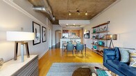 Located in Baltimore's vibrant Fell's Point neighborhood, this single-floor residence showcases a compelling blend of industrial and contemporary design. The open-concept living, dining, and kitchen area is anchored by striking exposed concrete ceilings with visible ductwork and track lighting, set above warm hardwood floors that create a bold contrast throughout the main living space. The kitchen is well-appointed with warm wood cabinetry, dark granite countertops, stainless steel appliances including a French door refrigerator and range, and a peninsula with bar seating — ideal for both cooking and casual dining. The living area offers generous space, complemented by industrial-style open shelving. The bedroom features plush carpeting, a concrete ceiling, and a large window framing city views, filling the room with natural light. The primary bathroom is finished with tile flooring, a soaking tub with tile surround, a long vanity with dark countertops, and access to a walk-in closet. Additional highlights include a dedicated office nook with carpet and a gallery wall, a full-size in-unit washer and dryer, and arched doorway details that add architectural character throughout the residence.