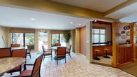 This thoughtfully designed single-story community space blends lodge-inspired architecture with warm, functional interiors throughout. The grand entrance foyer makes a strong first impression, featuring soaring vaulted ceilings clad in natural wood, a striking triangular clerestory window that floods the space with natural light, and a handcrafted wood staircase with vertical balusters leading to an upper level. A large potted tree anchors the transition between the tiled entry and adjoining spaces, adding a natural, organic element to the interior.

The lounge area is a standout feature, centered around a floor-to-ceiling stacked stone fireplace that serves as the architectural focal point of the room. Two upholstered armchairs flank the hearth, complemented by a patterned ottoman and side tables, creating an intimate seating arrangement bathed in natural light from nearby windows with wood-trimmed valances.

Adjacent to the lounge, a comfortable sitting room offers an orange sofa, accent chairs, a glass coffee table, and generous window coverage. The dining area features tile flooring, round pedestal tables with upholstered chairs in warm terracotta tones, and direct access to an outdoor patio. A bar counter with wooden stools provides an additional gathering point, completing this versatile and welcoming space.