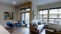 This single-floor space showcases a thoughtfully designed open-plan layout that seamlessly integrates the living and dining areas. The living area is anchored by a contemporary light gray sofa accented with patterned and green throw pillows, positioned to take advantage of natural light streaming through large windows. The windows offer views of the surrounding building exterior, filling the space with a bright, airy atmosphere.

The dining area features a round glass-top table paired with sleek blue upholstered chairs, providing an elegant yet functional setting for meals. Rich, dark hardwood-style flooring runs throughout, adding warmth and visual continuity to the open layout.

A standout feature is the full-height sliding glass door with transom windows above, which opens onto a private balcony equipped with outdoor seating — ideal for extending the living space outdoors. The neutral, warm-toned walls are complemented by modern recessed lighting overhead. Framed artwork adds a curated touch to the interior, while the overall design strikes a balance between contemporary style and comfortable functionality.