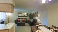 This single-floor residence offers a welcoming and functional layout designed for comfortable living. The open-concept floor plan seamlessly connects the living and dining areas, creating a sense of spaciousness and ease of flow throughout the space. The living area is anchored by plush, dark leather sofas accented with warm coral and patterned throw pillows, centered around a sturdy wooden coffee table that adds a rustic charm. A large decorative mirror with candle sconce accents adorns the wall, enhancing the sense of depth and reflecting light throughout the room. A lush indoor plant adds a natural, refreshing element to the corner of the living space. Adjacent to the living area, the dining space features a classic wooden table and chairs, setting the stage for casual meals or gatherings. Soft, neutral-toned carpeting runs throughout the main living areas, contributing to the warm and inviting ambiance. A textured ceiling adds architectural character overhead. The kitchen, visible through the open layout, provides convenient access and connectivity to the main living space. Overall, the residence offers a cozy, well-appointed environment with a harmonious blend of warm tones and functional design.
