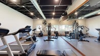 Located at 20 S Naper Blvd in Naperville, IL, this fitness center is a comprehensively equipped, single-floor facility designed to support a wide range of workout routines. The space features an exposed ceiling with dark-painted ductwork and structural elements, giving it an industrial aesthetic complemented by recessed and track lighting that keeps the area bright and functional. Natural light flows in through a row of windows along one wall, offering views of the surrounding greenery and contributing to an energizing atmosphere. A full wall of floor-to-ceiling mirrors visually expands the space and aids in proper form during workouts. The cardio section is well-stocked with treadmills, an elliptical trainer, and a stationary cycle, while the strength training area includes a multi-station cable machine and a dedicated weight bench. A separate functional training zone houses a tiered dumbbell rack, kettlebells, resistance bands, medicine balls, stability balls, and additional storage shelving. Large pedestal fans are positioned throughout for added ventilation and comfort. The rubber and hard flooring throughout is durable and suited for varied exercise activities. Posted fitness center regulations help ensure a safe and organized environment for all users.