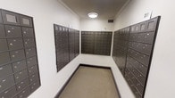 This compact, single-floor mail room is purpose-built to accommodate the postal needs of a multi-unit building. The room is lined on three sides with banks of dark bronze-finished individual mailboxes, offering a large number of secure, individually keyed units for residents. The mailboxes are arranged in neat rows and columns, maximizing wall space and ensuring organized mail distribution throughout the building. The room features clean white walls accented by crown molding near the ceiling, lending a polished, well-maintained appearance. A flush-mounted ceiling light provides consistent overhead illumination, keeping the space bright and functional. The flooring is covered in a neutral, low-pile carpet with a dark baseboard trim that frames the room neatly. A ventilation grille is visible near the ceiling, ensuring proper air circulation within the space. The overall design is straightforward and utilitarian, prioritizing accessibility and efficiency. The layout allows residents to move comfortably within the room while accessing their individual mailboxes, making it a practical and essential shared amenity within the building.