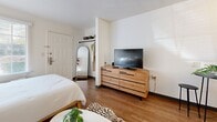 Located at 710 Yorkmont Rd in Charlotte, NC, this efficiently designed studio unit offers a thoughtfully arranged single-floor layout that maximizes every square foot. Warm wood-tone flooring runs throughout the space, lending continuity and a welcoming feel to the interior.

The open-concept living and sleeping area forms the heart of the unit, featuring a full-sized bed, a compact seating arrangement with a mustard-toned accent chair, and a wooden dresser with flatscreen TV. A large arched mirror and an open closet nook with overhead storage provide functional yet stylish organization. Colorful wall art adds personality to the crisp white walls.

The kitchenette is neatly tucked along one wall and includes white cabinetry with open wood-accented shelving, a full-size refrigerator, microwave, and a single-basin sink — offering all the essentials in a space-conscious footprint. A small bistro table with seating sits adjacent, providing a dedicated dining spot.

Track lighting on the ceiling ensures the space is well-lit, complementing the clean, modern aesthetic. The bathroom is a compact, functional space with white walls and standard fixtures. Overall, the unit presents a cohesive, urban studio aesthetic with practical amenities suited for comfortable everyday living in Charlotte.