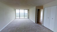 This single-floor residence offers a thoughtfully arranged layout centered around a generous open living area. The standout feature of the main living space is the expansive floor-to-ceiling window wall, which floods the room with abundant natural light and provides direct access to a private balcony. The balcony overlooks a lush, tree-covered landscape, offering a tranquil natural backdrop that enhances the overall ambiance of the space. The living area is finished with wall-to-wall carpeting that adds warmth underfoot, while a textured ceiling adds a classic architectural touch. Built-in shelving is conveniently integrated near the entryway to an adjoining room, providing ample storage and display space. The neutral color palette throughout — from the soft beige walls to the light carpeting — creates a calm, versatile environment well-suited to a variety of interior styles. The layout flows naturally from the main living area toward additional rooms, suggesting a practical and connected floor plan. Large sliding glass doors connect the interior to the balcony, blurring the boundary between indoor comfort and outdoor scenery, making this a space that balances functionality with a peaceful, light-filled atmosphere.