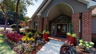 This single-story building presents a distinguished exterior defined by a combination of red brick accents and gray horizontal siding. The most prominent architectural feature is the sweeping arched portico above the main entrance, which frames double wood-and-glass doors topped by a decorative arched transom window that draws the eye upward and introduces natural light into the entryway. Brick columns flank the entrance, reinforcing the traditional aesthetic and providing structural definition to the covered porch area.

The surrounding landscaping is carefully maintained, with curved concrete walkways leading to the entrance through beds of colorful flowering plants, ornamental shrubs, and mulched ground cover. Bold red planters filled with lush greenery provide striking visual accents on either side of the entry doors. Mature trees in the background contribute to a well-established, park-like setting.

The exterior brick walling features detailed coursework, visible at the building's base and along its corners, demonstrating quality craftsmanship throughout the construction. A covered side entrance with a concrete porch and a second blue door offers additional access to the building. The overall exterior presents a polished, professional appearance well-suited for a community or commercial facility.