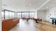 This thoughtfully designed communal space combines a relaxed lounge area with a well-appointed dining and gathering zone, all on a single floor. The interior is defined by a wall of large, floor-to-ceiling windows framed in dark metal, flooding the space with abundant natural light and offering panoramic views of the surrounding cityscape and treetops beyond.

The flooring throughout is a warm, wide-plank hardwood laid in a chevron-inspired pattern, lending the space a refined yet welcoming character. The lounge area is anchored by a pair of rich cognac leather Chesterfield sofas arranged around a dark wood coffee table, accompanied by wall-mounted sconces and a flat-screen television — creating an inviting retreat for relaxed gatherings. A door leads to an outdoor terrace, further extending the connection to the outdoors.

The dining area features multiple solid wood tables paired with distinctive bentwood chairs and upholstered bench seating, offering flexible arrangements for both intimate and group settings. Pendant lights hang above the tables, complementing the recessed lighting overhead and adding warmth to the bright, airy atmosphere.

The clean white walls and crisp ceilings maintain a modern aesthetic throughout, while the blend of leather, wood, and natural light creates a space that is both functional and stylishly comfortable.