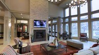 This elegant communal lounge is designed to impress from the moment you enter. The centerpiece of the space is a striking floor-to-ceiling stacked stone fireplace, topped with a wall-mounted flat-screen television, creating a compelling focal point that anchors the entire room. The soaring double-height ceilings lend an airy, grand atmosphere, further enhanced by a stylish chandelier that adds a touch of sophistication overhead.

Natural light pours in generously through expansive floor-to-ceiling windows and glass doors along the exterior wall, offering pleasant views of the surrounding outdoor area. Neutral-toned drapery frames the windows, adding a refined finishing touch. The lounge is thoughtfully furnished with a mix of plush seating arrangements, including sofas, accent chairs with bold patterned upholstery, and a sturdy rectangular coffee table, all set atop a rich, warm-toned area rug.

Multiple seating clusters throughout the space encourage both intimate conversation and larger group gatherings. Potted greenery is placed strategically to introduce a fresh, natural element. The overall ambiance is warm, inviting, and well-appointed — a comfortable and visually dynamic space suited for relaxation and social interaction.