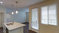 This single-floor residence showcases a clean, contemporary design with an open-concept layout that seamlessly connects the kitchen and living areas. The kitchen is a standout feature, centered around a sleek white island with an integrated stainless steel sink and ample counter space, making it ideal for both meal preparation and casual dining. White cabinetry lines the walls, providing generous storage, while stainless steel appliances, including a refrigerator and gas range, add a modern, professional touch. Overhead, recessed lighting is complemented by elegant pendant lights suspended above the island, creating a warm and functional ambiance. Dark hardwood-style flooring runs throughout, providing a rich contrast to the crisp white walls and cabinetry. The living area flows naturally from the kitchen, offering a versatile open space bathed in natural light from a full-length glass door with sidelights fitted with white blinds, hinting at outdoor access. A hallway leads to additional areas of the unit, including built-in storage. The overall aesthetic is bright, minimalist, and move-in ready.