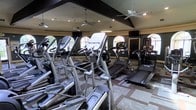 This fitness center offers a comprehensive workout environment designed to meet a variety of exercise needs. The space is anchored by a generous selection of cardio equipment, including multiple elliptical trainers, treadmills, and additional exercise machines arranged in organized rows for easy access. The room's architectural design features vaulted ceilings accented by dark exposed beams, which lend a sense of height and openness to the interior. Multiple ceiling fans are mounted throughout, ensuring consistent air circulation during workouts. Large arched windows line the walls, allowing ample natural light to filter in while providing views of the surrounding exterior. The walls are finished in a warm, neutral tone complemented by dark trim details, creating an inviting yet focused atmosphere. Mounted flat-screen televisions are positioned throughout the room, offering entertainment during exercise sessions. The flooring features a patterned tile design that adds a decorative touch while remaining practical for a high-traffic fitness environment. Overall, this fitness center combines functional equipment with thoughtful architectural details to create a comfortable and motivating space for exercise.