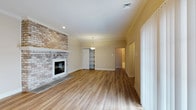 Located at 1 Marlbrook Lane in Lansdale, Pennsylvania, this single-floor residence offers a thoughtfully designed layout with a warm and inviting ambiance throughout. The main living area immediately draws attention with its striking floor-to-ceiling brick accent wall framing a classic fireplace, serving as a bold architectural centerpiece. Rich, warm-toned hardwood floors extend throughout the space, lending a sense of continuity and elegance to the interior. Crown molding adds a refined finishing touch along the ceiling line, complementing the neutral, cream-toned walls that keep the space feeling bright and open. A pendant light fixture provides a subtle focal point, while recessed lighting ensures the room is well-illuminated. A built-in shelving unit offers functional storage and display space, adding character to the interior. Wide vertical blinds on the windows allow for flexible light control, contributing to the airy feel of the living area. A hallway leads further into the home, hinting at additional private spaces beyond. The overall design balances classic architectural details with practical, everyday functionality, making this a comfortable and well-appointed single-level home in the heart of Lansdale, PA.
