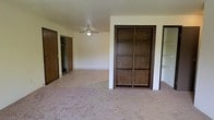 This single-floor space presents a practical and functional layout designed to maximize storage and usability. The main living area features a combination of plush carpeting and smooth tile flooring, creating a clear distinction between zones within the open layout. A ceiling fan with an integrated light fixture is centrally positioned, providing both air circulation and ambient lighting to the space.

A standout built-in shelving unit with dark wood framing serves as a prominent architectural feature, offering multiple open shelves ideal for display or organization. The warm wood tones of the shelving add a classic contrast to the neutral, light-colored walls throughout.

Multiple closets are accessible from the main area, including what appears to be a walk-in closet and additional standard closets, providing ample storage solutions. The doors feature matching dark wood finishes that complement the built-in shelving, contributing to a cohesive interior aesthetic.

The space is finished with clean, white walls that reflect light and contribute to an open, airy feel. Overall, the layout is straightforward and versatile, well-suited for a variety of furniture arrangements and personal configurations.