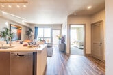 This well-appointed single-floor residence offers a thoughtfully designed open-concept layout that seamlessly connects the kitchen, dining, and living areas. The kitchen is a standout feature, showcasing rich dark-wood cabinetry, light quartz countertops, a decorative tile backsplash, stainless steel appliances including a refrigerator, range, and microwave, and a large center island with bar seating — ideal for both meal preparation and casual dining. Track lighting overhead ensures excellent illumination throughout the cooking space.

The living area is bright and inviting, anchored by large windows that invite abundant natural light and offer open outdoor views, with access to a balcony. Wide-plank wood-style flooring flows throughout the main living spaces, adding warmth and continuity to the design.

The primary bedroom is generously sized, featuring large windows with scenic views, plush carpeting, and a dedicated walk-in closet. A secondary bedroom also includes a walk-in closet for added storage convenience.

The full bathroom is finished with a dark wood vanity, a wide mirror, and a combination tub and shower. The neutral gray palette and wood-tone accents create a cohesive, contemporary aesthetic that carries throughout the entire residence.