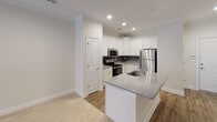 This single-floor residence offers a thoughtfully designed layout that balances comfort and functionality. The open-concept kitchen serves as a central highlight, featuring crisp white shaker cabinetry, granite countertops, a dark subway tile backsplash, and a full suite of stainless steel appliances including a range, over-the-range microwave, and refrigerator. A peninsula with an integrated sink provides additional workspace and casual seating, seamlessly connecting the kitchen to the adjacent living area. The living room is finished with plush carpeting, crown molding, a ceiling fan with light fixture, and French doors that invite natural light and provide access to an outdoor area. The bedrooms continue the warm, neutral palette with soft carpeting, ceiling fans, and windows dressed with blinds. One bedroom includes direct access to a walk-in closet with built-in shelving, offering practical storage. The bathroom is finished with wood-look flooring, a tub-shower combination with white tile surround, and brushed nickel fixtures and hardware. Throughout the residence, consistent warm-toned walls, white trim, and wood-look hard flooring in the main areas create a cohesive and inviting atmosphere.