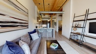 Located at 165 Thorndike, this single-floor loft-style residence showcases a compelling blend of industrial heritage and contemporary design. The open-concept living and kitchen area immediately impresses with soaring wood-plank ceilings, exposed structural columns, and visible ductwork that nod to the building's historic character. Light wood-tone flooring runs throughout, unifying the space and complementing the warm tones overhead.

The kitchen is well-appointed with wood-grain cabinetry, stainless steel appliances, a white tile backsplash, and a peninsula with bar seating — ideal for casual dining or entertaining. Pendant lighting drops from the dramatic ceiling, adding warmth to the workspace. The living area benefits from large windows flanked by original exposed brick, allowing generous natural light to pour in and providing an authentic industrial focal point.

The bedrooms are calm and well-proportioned, with high ceilings continuing the sense of openness. The bathroom features marble-surround tile on a tub-shower combination enclosed by a glass panel, lending a clean, upscale feel. A dedicated laundry area with a stacked washer and dryer adds everyday convenience. Throughout, the design balances raw architectural elements with refined modern finishes.