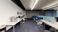 Located at 29 E Green St in Champaign, Illinois, this single-floor commercial workspace is thoughtfully designed to support a range of work styles and collaborative needs. The layout balances open and enclosed areas to create a dynamic, functional environment throughout.

The open workspace area features rows of sleek white workstations with blue ergonomic chairs and teal privacy dividers, providing an organized setting for focused individual work. A dedicated computer lab station with desktop computers and printing equipment is conveniently integrated into this zone.

Glass-walled private offices line one section of the space, offering visual transparency while maintaining acoustic separation. Each enclosed room is equipped with a conference table and chairs, with large windows providing views of the surrounding streetscape and natural light filtering through roller shades.

A welcoming lounge area near the exterior-facing windows serves as a relaxed gathering zone, furnished with modular sofas and lounge chairs in neutral tones alongside low coffee tables. Geometric-patterned carpet tiles in shades of grey, blue, and gold add visual interest underfoot throughout the space.

Near the entrance, a casual seating nook with navy upholstered armchairs and a round table provides an informal meeting spot. The exposed black ceiling beams contrast with the white walls, giving the space a contemporary, urban aesthetic. Overall, the design promotes collaboration, productivity, and comfort in equal measure.